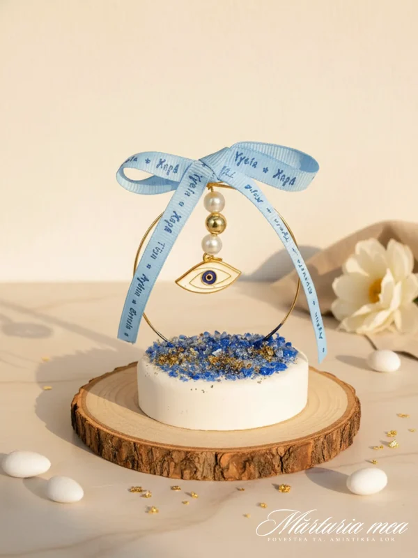 Marturie cu Simbol Ochi Stilizat – Cadou Elegant de Protecție pentru Botez și Nuntă | Marturia Mea marturie simbol ochi protectie pe baza rotunda alba cu pietricele decorative albastre si auriu si fundita bleu