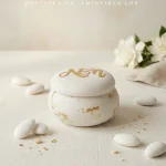Cutiuță elegantă pentru mărturii în formă de macaron alb, închisă, personalizată pe capac cu inițiale aurii, așezată pe un fundal luminos alături de bomboane albe glazurate.